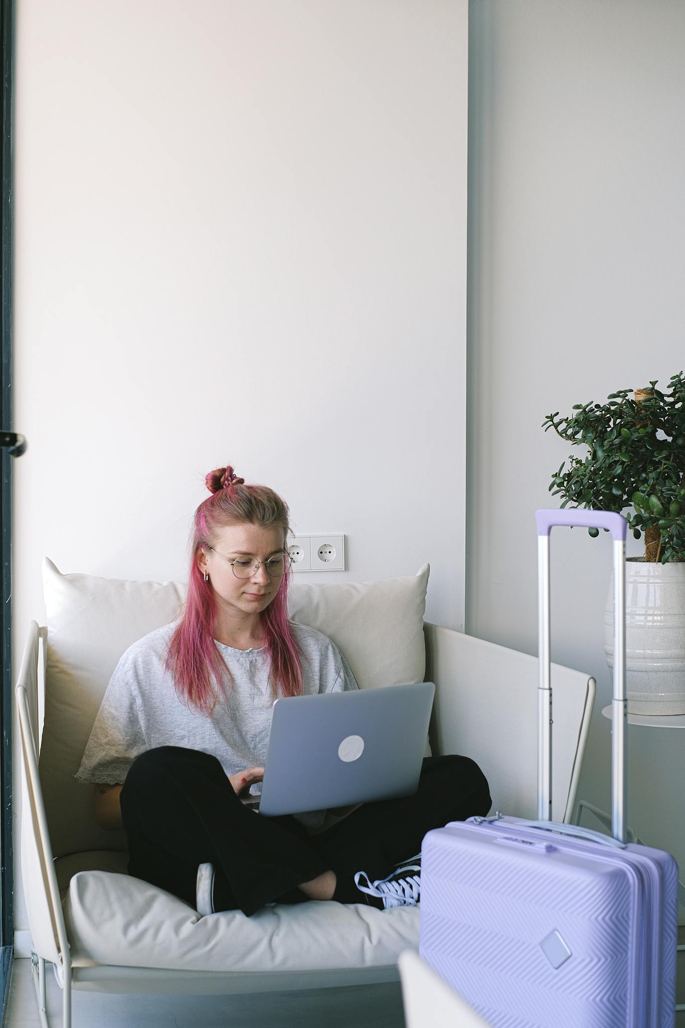 Woman with pink hair working on a laptop while sitting on a sofa in a hotel room with luggage.